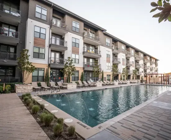 A modern four-story HOA-managed apartment community with a long resort-style swimming pool, lounge chairs, paver walkways, manicured plantings, and architectural facades in late-day light. The kind of maintenance property a landscape account manager walks during a SiteDNA pre-inspection before submitting a competitive bid to the HOA board.